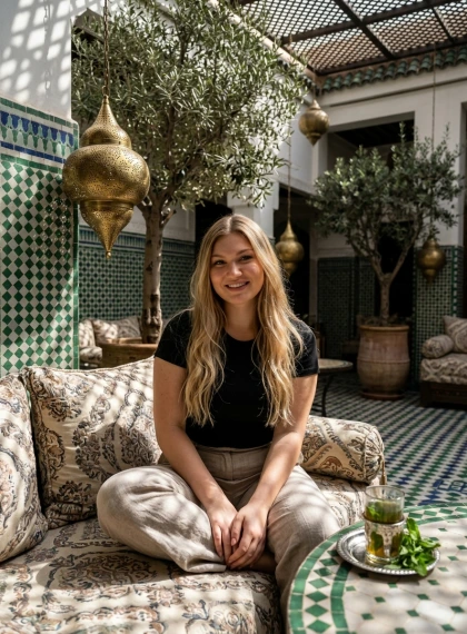Sitting cross-legged on shaded lounge seating in a Marrakech riad courtyard, surrounded by zellige tile walls and oli...