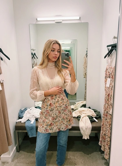 Spontaneous mirror selfie in a boutique fitting room, one hand holding phone at angle, other smoothing a patterned dr...